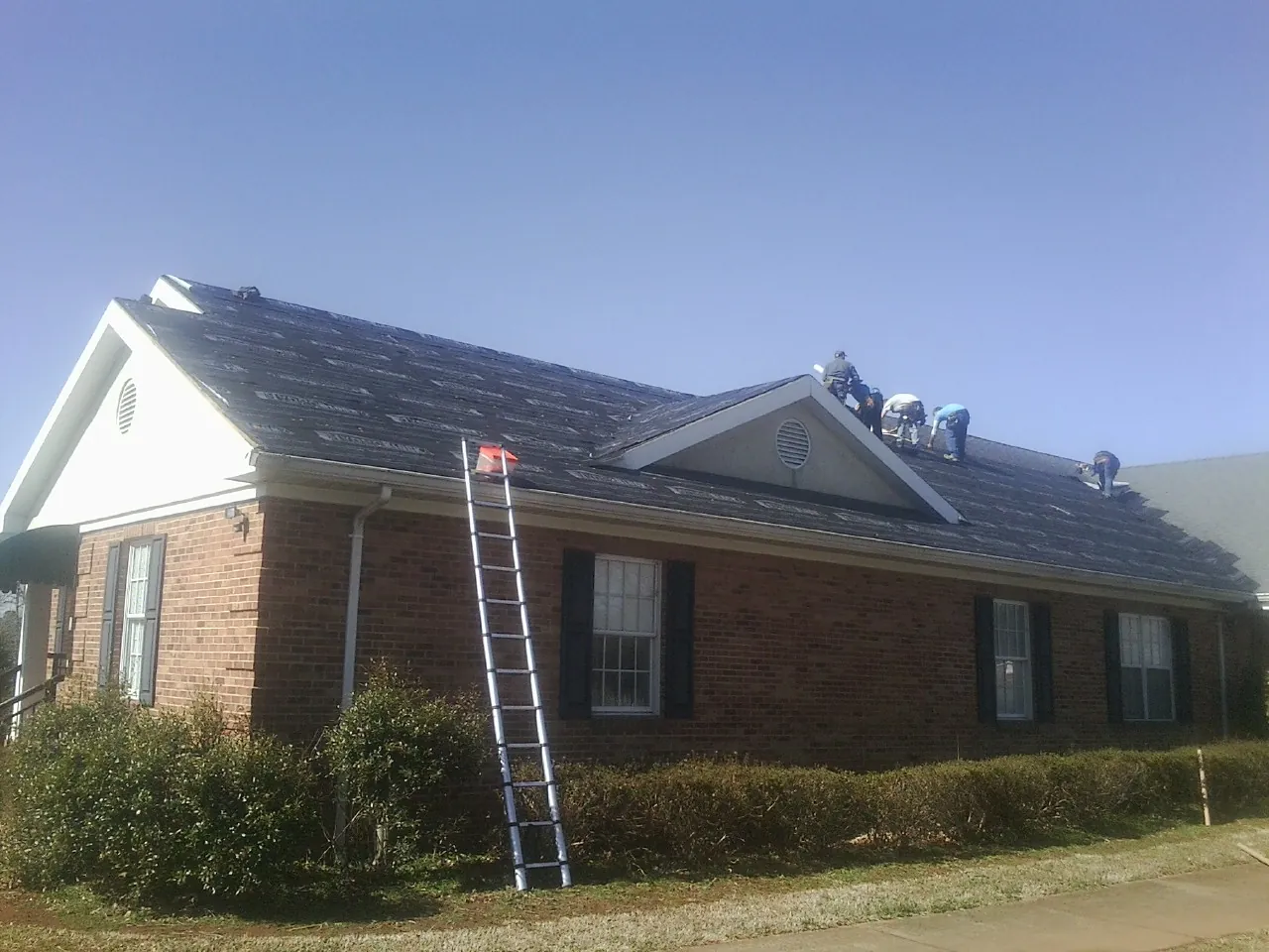 Crew tearing shingles off a brick ranch roof with a ladder set — Emergency Roof Repair work by our crew in Buffalo Center