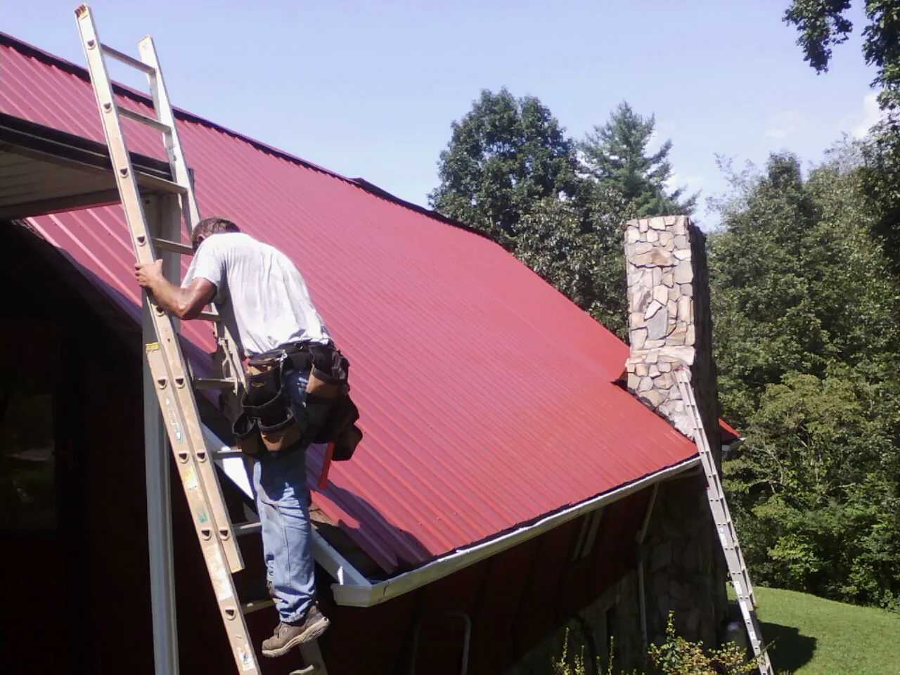 Roofer on a ladder beside a red metal roof and stone chimney — Soffit & Fascia Repair by Roofing Brothers in Buffalo Center